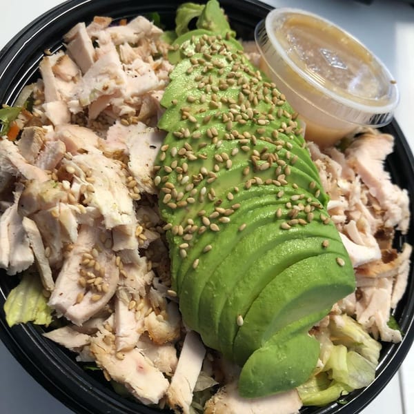 A Blue Fin chicken salad with sliced avocado, sesame seeds, and dressing in a black bowl.