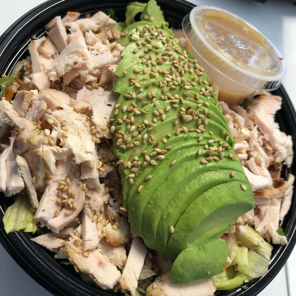 A Blue Fin chicken salad with sliced avocado, sesame seeds, and dressing in a black bowl.